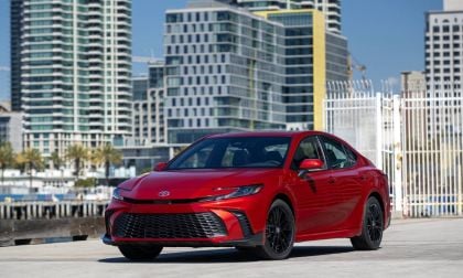 A sleek red Toyota sedan parked in an urban setting with modern skyscrapers and palm trees in the background.