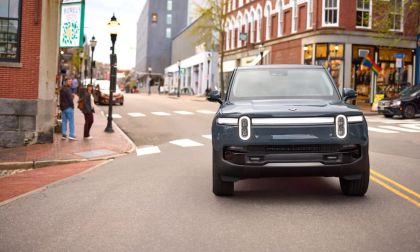 Front view of a Rivian R1S electric SUV driving through an urban downtown street, featuring signature vertical LED headlights and a boxy adventure-focused design.