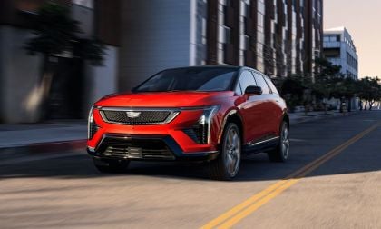 Red SUV driving down a city street with buildings and trees in the background.