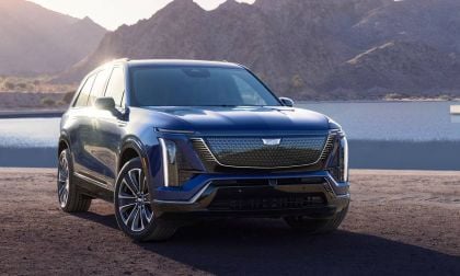A sleek, dark blue 2026 Cadillac Vistiq parked by a serene lake, with mountains in the background and a sailboat on the water.