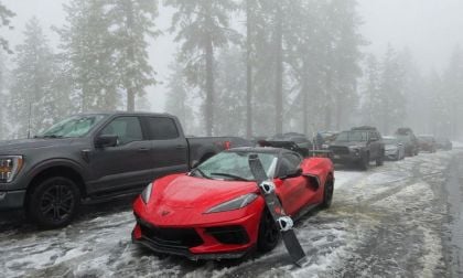 Red Chevrolet Corvette C8 parked in a snowy mountain lot with a pair of skis leaning on the front bumper, surrounded by snow-covered trucks and trees.