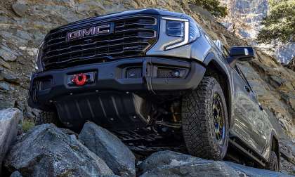 A grey 2026 GMC Sierra 1500 AT4 truck is posted up on rocky terrain, emphasizing rugged durability. The truck's front is prominent, with a logo and skid plate visible.