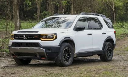 2026 Honda Pilot TrailSport in Platinum White Pearl, front three-quarter view, featuring black wheels, all-terrain tires, and rugged styling, parked on dirt trail in wooded setting.