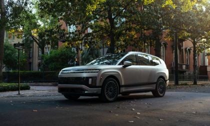A silver Hyundai Ioniq 9, viewed from a front three-quarter angle, parked on a tree-lined historic city street with brick buildings in the background.
