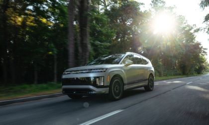 Silver SUV driving on tree-lined road with sunlight streaming through forest canopy