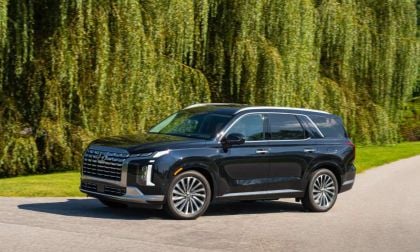 2026 Hyundai Palisade in black, photographed at three-quarter front angle on paved road beneath weeping willow trees, featuring distinctive split LED headlights and large grille.