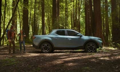 A light blue Hyundai Santa Cruz is parked in a sunlit forest. Two people with backpacks talk nearby, surrounded by tall trees, conveying a sense of adventure.