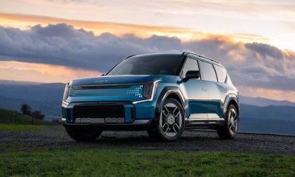 Blue Kia EV9 SUV parked on hillside against dramatic sunset sky with mountains.