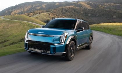 A blue 2026 Kia EV9 with modern styling drives on a winding road through green hills and mountains, conveying a sense of adventure and freedom.