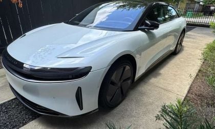 A sleek white electric car with black accents parked on a concrete driveway, surrounded by greenery and a dark wooden fence.