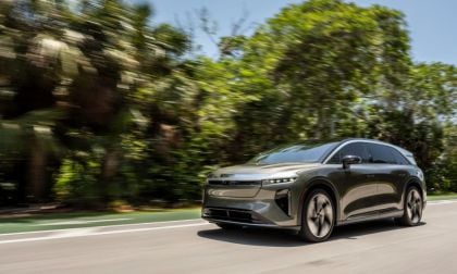 2026 Lucid Gravity shown in three-quarter front view, driving on tree-lined road with distinctive LED lighting and aerodynamic wheels.