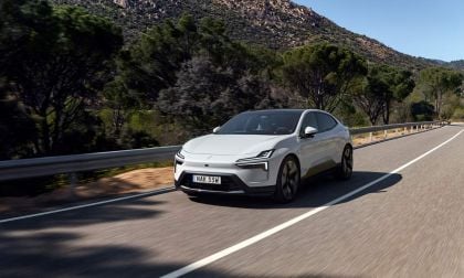 A sleek, white electric car drives along a winding road, surrounded by lush trees and a mountainous backdrop under clear skies.