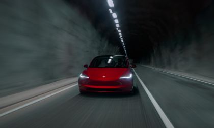 A red Tesla Model 3 shown from the front three-quarter angle driving through a dimly lit tunnel, with its distinctive LED headlights illuminated and creating a dramatic atmosphere.