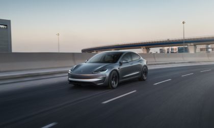 A gray electric car drives on a highway near an overpass at dusk.