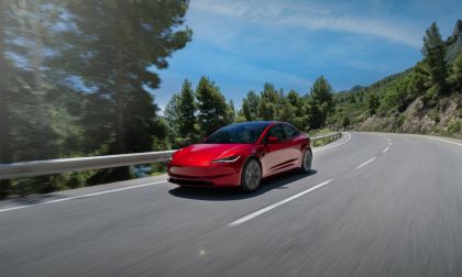 Candy red Tesla Model 3 driving on a winding mountain road, photographed from front three-quarter view, surrounded by pine trees and guardrails.