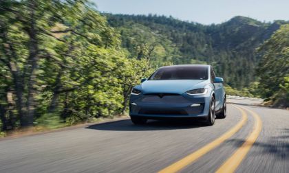 A light blue Tesla Model X SUV shown from a front three-quarter angle, driving on a winding mountain road surrounded by lush green forest.