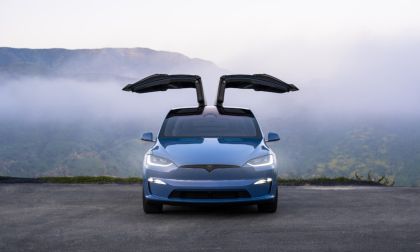 Blue Tesla Model X with signature falcon-wing doors open, front view, parked on mountain overlook with misty valley background.