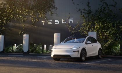 White Tesla Model Y parked at a Tesla Supercharger station with Tesla logo signage in the background.