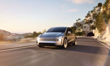 Photograph of a 2026 Tesla Model Y electric vehicle in the Juniper color, captured from a front three-quarter angle view as it drives on a road in California surrounded by lush greenery and scenery.