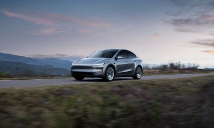 Silver 2026 Tesla Model Y electric SUV photographed from front three-quarter angle, driving on mountain road at dusk with dramatic cloudy sky.