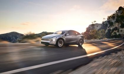 Photograph of a 2026 Tesla Model Y electric vehicle in the Juniper Silver color, captured from a three-quarter front angle with a sunset landscape visible in the background.