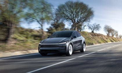 Electric car driving on a rural road with trees and clear sky in the background.