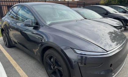 Gray Tesla Model Y Juniper, photographed at three-quarter front angle in parking lot, featuring sleek aerodynamic design, black wheels, and glass panoramic roof.