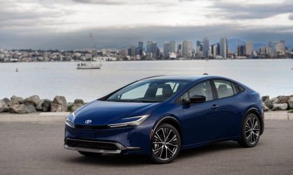 A blue Toyota Prius parked by the waterfront, with a city skyline and cloudy sky in the background.