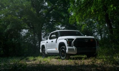 A white 2026 Toyota Tundra pickup truck is shown from a front three-quarter angle, parked in a wooded setting with lush green trees in the background.
