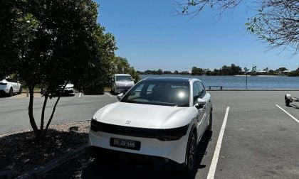 White car parked in a lot near a lake, surrounded by trees and other vehicles.