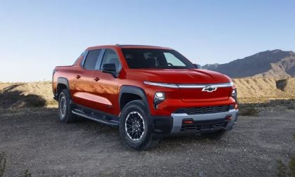 2026 Chevrolet Silverado EV electric pickup truck in red, front three-quarter view off-roading on desert terrain with rugged tires and bold grille design.