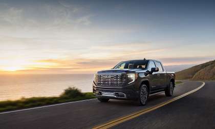 2026 GMC Sierra Denali Ultimate driving on a coastal road at sunset, shown from the front three-quarter angle.
