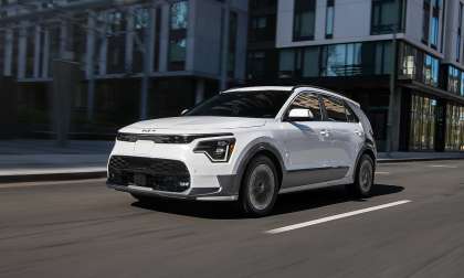 White 2026 Kia Niro EV driving through a city street in a front three-quarter action view.