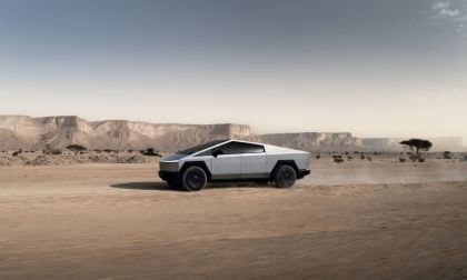 A silver Tesla Cybertruck shown in side profile view against a desert landscape with rocky cliffs and scattered trees in the background.
