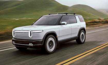 White 2027 Rivian R2 driving on a two-lane road through green hills in a front three-quarter action view.