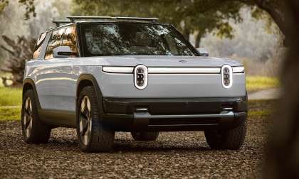 White 2027 Rivian R2 parked on a wooded trail in a front three-quarter view.