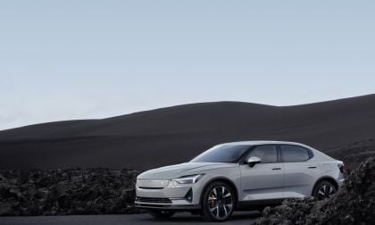 A white Polestar 2 parked on a barren landscape, with dark volcanic soil and a soft blue sky in the background.