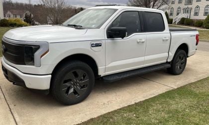 A white Ford F-150 Lightning truck parked on a driveway, showcasing its sleek design and black alloy wheels.