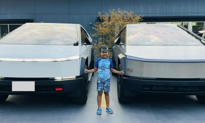 A child smiles between two Tesla Cybertrucks with metallic finishes. The scene is calm and set against a modern building with plants in the background.