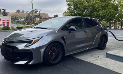 A polished gray Toyota GR Corolla is parked at a gas station while fueling up. It features tinted windows and red brake calipers with trees in the background.