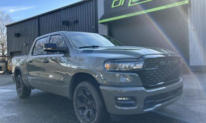 A gray 2026 Ram 1500 pickup truck shown from a front three-quarter angle, featuring black wheels and the aggressive RAM grille design, parked outside a building with a green sign.