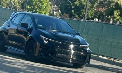 Black Toyota hatchback parked on asphalt with green fence and trees in background