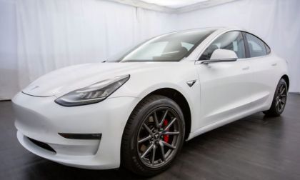 A white Tesla Model 3 with a modern design is parked indoors on a showroom floor. It features aerodynamic curves, sharp headlights, and distinctive black wheels.