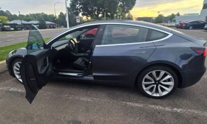 A dark grey Tesla Model 3 is parked with the driver’s door open under a cloudy sky. The setting is a quiet parking lot, evoking tranquility.