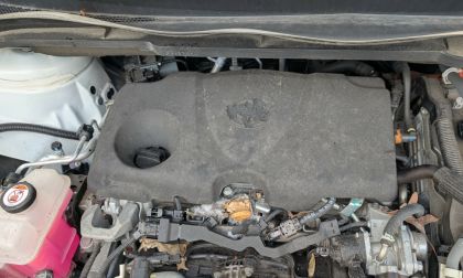 Anna's 2026 Toyota Sienna's bay with a dusty engine cover is shown. A worn emblem is visible on the cover. Loose wires and a pink coolant reservoir are noticeable nearby.