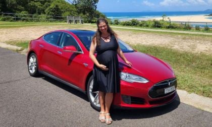 Anne Bishop in a black dress smiles while standing besides her red Tesla Model S on a scenic road. The background features greenery and a view of the ocean under a clear sky.