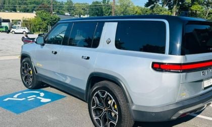 A gray Rivian R1S with tinted windows is parked in a handicap spot. The electric vehicle's sleek design and modern features are visible under a sunny sky.