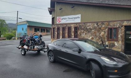 A black Tesla Model 3 is parked at a three-quarter front angle outside eMoto Powersports, with motorcycles on a trailer behind it.