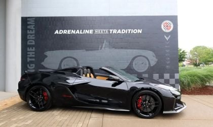 A sleek black Corvette Z06 convertible parked beside a mural showcasing classic Corvettes and text reading "ADRENALINE MEETS TRADITION."