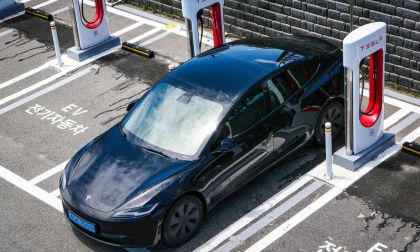 A black Tesla electric vehicle parked at a charging station, with wet pavement and marked EV parking spaces.